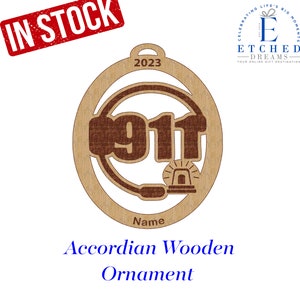 May include: A brown wooden ornament shaped like a circle with the number 911 inside. The ornament has a headset and a siren on it. The text "2023" is at the top of the ornament. The text "Name" is at the bottom of the ornament. The text "Accordian Wooden Ornament" is below the ornament.