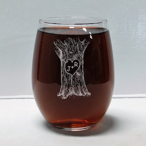 May include: A clear glass wine glass with a design etched into the side. The design is of a tree with a heart carved into the trunk. The heart has the initials "J+R" inside. The glass is filled with red wine.