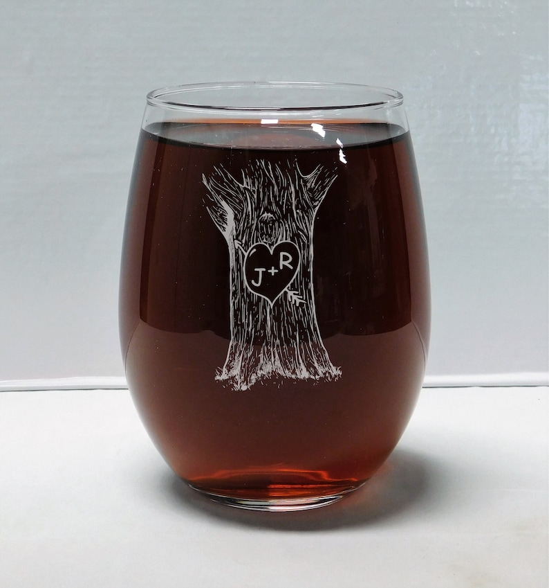 May include: A clear glass wine glass with a design etched into the side. The design is of a tree with a heart carved into the trunk. The heart has the initials "J+R" inside. The glass is filled with red wine.