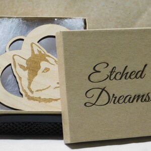 May include: A wooden ornament in the shape of a heart with a husky dog's head inside. The ornament is in a brown box with the words "Etched Dreams" on the front. The ornament also has the year "2018" engraved on it.