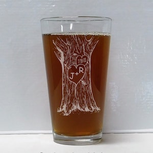 May include: A clear glass pint glass with a brown liquid inside. The glass has an etched design of a tree with a heart carved into it. The heart has the initials "J+R" and the date "7/18/20" etched into it.