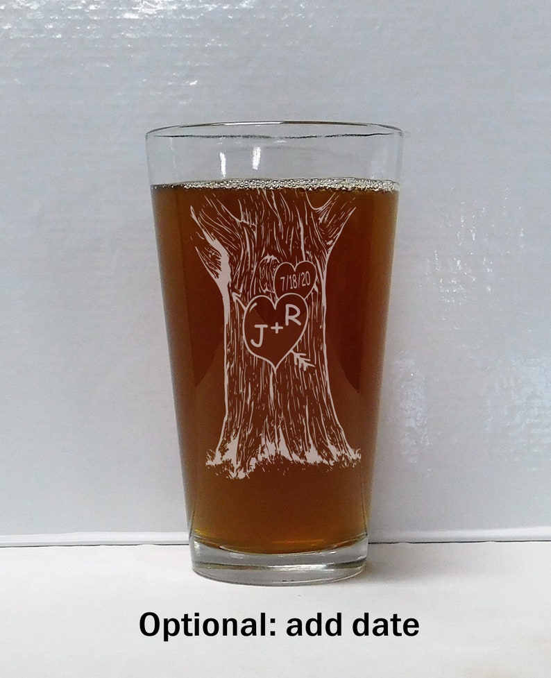 May include: A clear glass pint glass with a brown liquid inside. The glass has an etched design of a tree with a heart carved into it. The heart has the initials "J+R" and the date "7/18/20" etched into it.