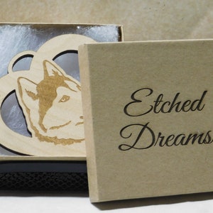 May include: A wooden ornament in the shape of a heart with a husky dog's head inside. The ornament is in a brown box with the words "Etched Dreams" on the front. The ornament also has the year "2018" engraved on it.