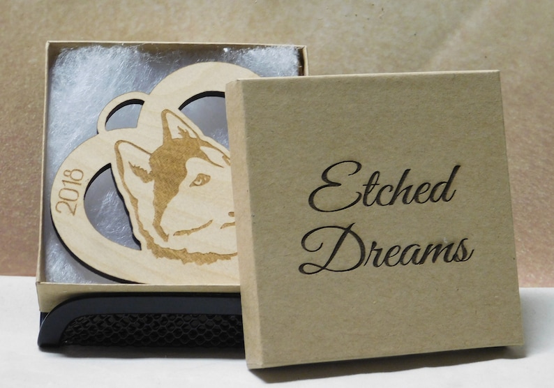 May include: A wooden ornament in the shape of a heart with a husky dog's head inside. The ornament is in a brown box with the words "Etched Dreams" on the front. The ornament also has the year "2018" engraved on it.