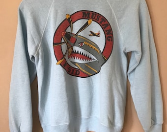 May include: Light blue sweatshirt with a graphic of a fighter plane with the words "Mustang" and "P-51D" in a red circle.