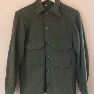 Puede incluir: Una camisa de lana verde con cierre de botones y dos bolsillos grandes. La camisa tiene un estilo clásico de trabajo.