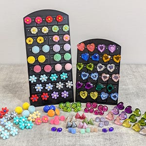 May include: Assortment of colorful earrings displayed on black earring stands. The earrings are in various shapes, including flowers, hearts, and geometric shapes. The earrings are made of plastic and metal.