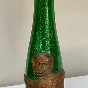 May include: Green glass bottle with a glitter finish and a brown monkey figure wrapped around it. The bottle has a long neck and a rounded base. Decorative item.