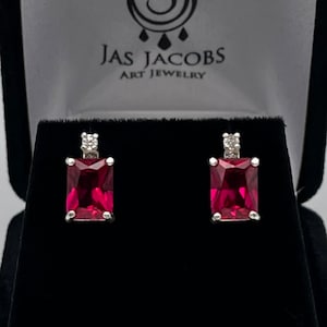 May include: A pair of ruby-colored, rectangular-cut gemstone earrings set in silver. Each earring features a small, square, clear stone at the top. The earrings are displayed in a black jewelry box with the text "Jas Jacobs Art Jewelry".