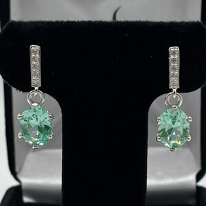 May include: A pair of silver dangle earrings with a light green gemstone and a row of clear stones on the top of the earring.