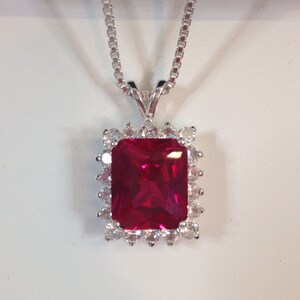 May include: A silver necklace with a rectangular, ruby-red pendant. The pendant is surrounded by a halo of smaller, clear gemstones. The chain is a delicate box chain style, and the pendant is centered on a white background.