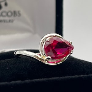 Puede incluir: Un anillo de plata con una piedra preciosa de rubí en forma de pera. El anillo se muestra en una caja de terciopelo negro.