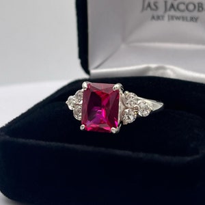 May include: A silver ring with a large, square-shaped, bright pink gemstone surrounded by smaller, clear gemstones. The ring is in a black velvet box with the words "Jas Jacob Art Jewelry" printed on the inside.