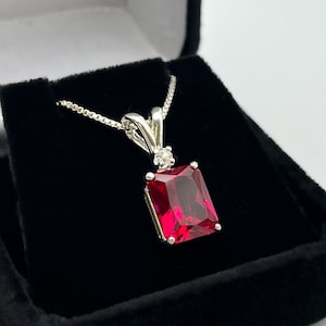 Puede incluir: Un collar de plata con un colgante de piedra roja y un pequeño acento de piedra clara. El collar está en una caja de terciopelo negro con las palabras "Jas Jacobs Art Jewelry" en la tapa.