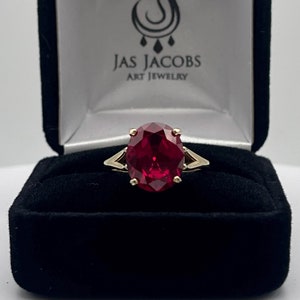 May include: A red gemstone ring with a gold band. The ring is in a black velvet box with the words "Jas Jacobs Art Jewelry" on the lid.