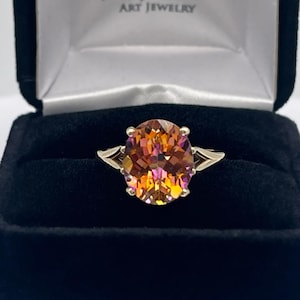 May include: A gold ring with a large, round, orange and pink gemstone. The ring is in a black velvet box with the words "Jas Jacobs Art Jewelry" on the lid.