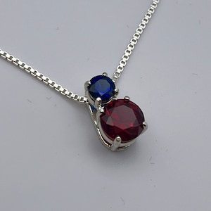 May include: A silver necklace with a pendant featuring two gemstones. The top gemstone is a deep blue, and the bottom is a vibrant red. The chain is a delicate, square-link style, and the pendant is set in a silver metal.