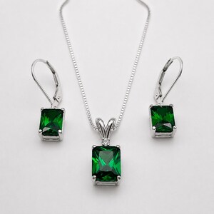 May include: A silver jewelry set consisting of a pendant necklace and two matching leverback earrings. Each piece features a rectangular emerald green gemstone set in a silver frame, with a small clear stone accent above the main gem.