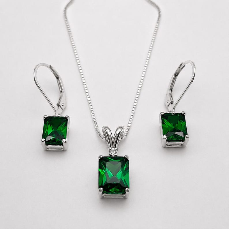 May include: A silver jewelry set consisting of a pendant necklace and two matching leverback earrings. Each piece features a rectangular emerald green gemstone set in a silver frame, with a small clear stone accent above the main gem.