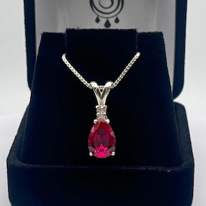 May include: A silver pendant necklace with a pear-shaped, deep red gemstone. The pendant is set in a silver setting with a small accent stone above the main gem. The necklace is displayed in a black jewelry box.