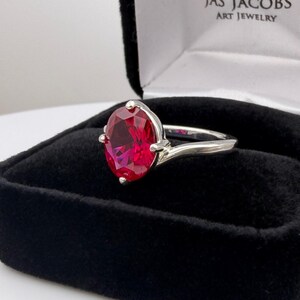 May include: A silver ring with a large, oval-cut, deep red gemstone. The ring is set in a classic style, with the gemstone held in place by four prongs. The ring is displayed in a black velvet jewelry box.