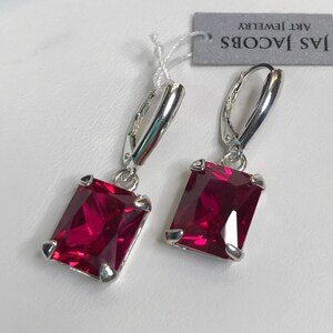 May include: A pair of silver-toned drop earrings featuring rectangular, deep red gemstones. The earrings have a lever-back closure and are labeled "JAS JACOBS ART JEWELRY". The gemstones are set in a silver-toned metal.