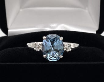 May include: A silver-toned ring featuring a large, oval-cut, light blue center stone held by four prongs. The band includes a smaller, round-cut clear stone on each side of the center setting. The ring is presented in a black jewelry box.