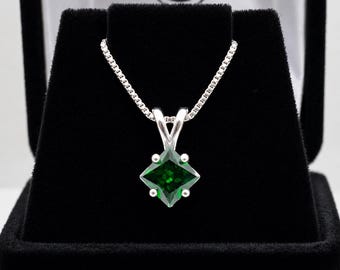 May include: A silver necklace featuring a square-cut, deep green gemstone pendant held by a four-prong setting. The pendant hangs from a delicate silver box chain, presented against a black velvet display stand with a white interior lining.