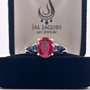 May include: A silver ring with a large oval-cut red gemstone, flanked by two smaller pear-shaped blue gemstones. The ring is displayed in a black velvet jewelry box. The box has a white interior with the text "JAS JACOBS ART JEWELRY".