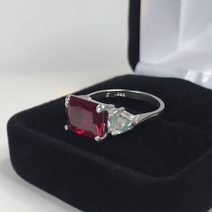 May include: A silver ring with a large, rectangular, red gemstone and two smaller, clear gemstones. The ring is in a black velvet ring box.