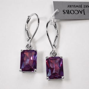Peut inclure: Une paire de boucles d'oreilles à fermoir levier argenté avec des pierres rectangulaires à facettes violettes. Une étiquette grise est attachée à une boucle, portant le texte JACOBS ART JEWELRY en noir. Les boucles ont une finition métallique polie.