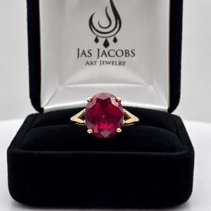 May include: A gold ring featuring a large, oval-cut, deep red gemstone held by four prongs. The ring is displayed in a black velvet box with the text JAS JACOBS ART JEWELRY printed in black on the white interior lid lining.