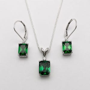 May include: A silver jewelry set consisting of a necklace and two leverback earrings. Each piece features a rectangular emerald-green gemstone. The pendant is accented with a small clear stone at the top, suspended from a delicate silver chain.
