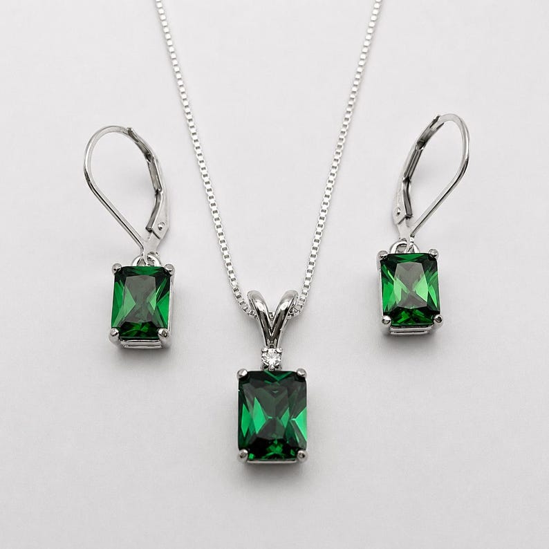 May include: A silver jewelry set consisting of a necklace and two leverback earrings. Each piece features a rectangular emerald-green gemstone. The pendant is accented with a small clear stone at the top, suspended from a delicate silver chain.