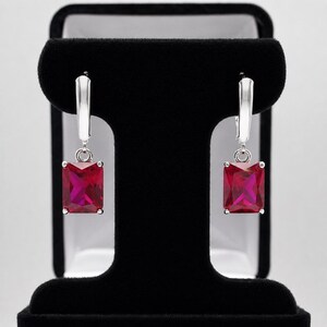 May include: A pair of silver-toned drop earrings featuring rectangular, faceted, deep red stones. Each earring has a polished metal lever-back closure attached to a small jump ring that connects to the vibrant, rectangular red gemstone pendant.