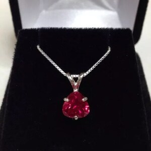 May include: A silver necklace featuring a heart-shaped, vibrant red pendant, set against a black velvet background. The pendant is a faceted gemstone. The necklace is presented in a jewellery box.