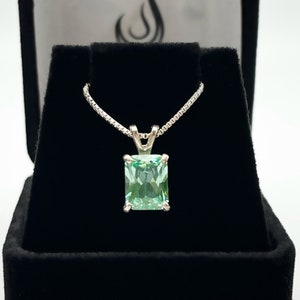 May include: A silver chain necklace with a light green emerald-cut gemstone pendant. The pendant is set in a silver bezel setting and is displayed in a black velvet box.