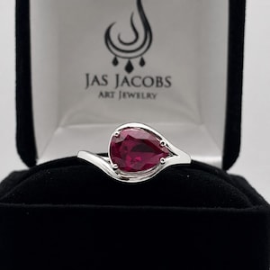 May include: A silver ring featuring a pear-shaped, deep red gemstone set in a bypass-style band. The ring is displayed inside a black velvet jewelry box with the text JAS JACOBS ART JEWELRY printed in black on the white interior lid lining.