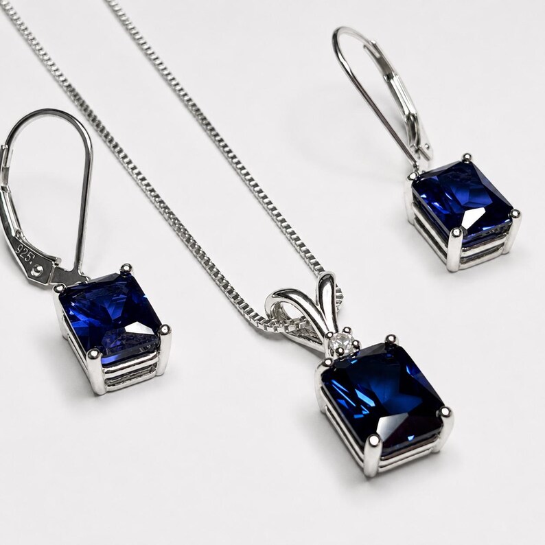May include: A jewelry set consisting of a silver necklace and two matching leverback earrings. Each piece features a rectangular, deep blue gemstone set in a silver frame. The necklace includes a thin silver chain and a small clear accent stone.