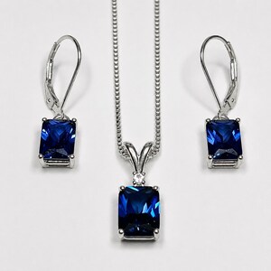 May include: A jewelry set consisting of a silver-toned necklace and two matching lever-back earrings. Each piece features a rectangular, deep blue faceted gemstone set in a silver-toned metal frame, with a small clear stone above the pendant.