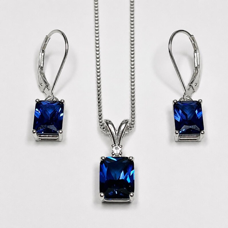 May include: A jewelry set consisting of a silver-toned necklace and two matching lever-back earrings. Each piece features a rectangular, deep blue faceted gemstone set in a silver-toned metal frame, with a small clear stone above the pendant.