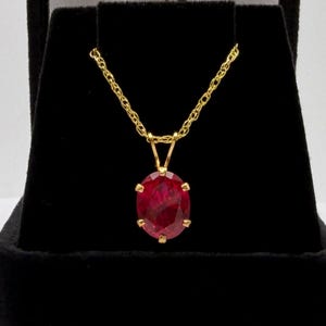 May include: A gold necklace with a ruby pendant. The pendant is an oval-shaped, deep red gemstone set in a gold bezel. The chain is a delicate gold cable chain, displayed against a black background.