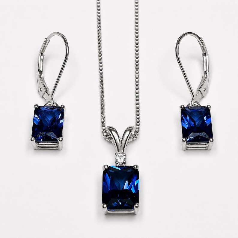 May include: A jewelry set consisting of a silver-toned necklace and two matching earrings. Each piece features a rectangular, deep blue faceted gemstone set in a silver-toned frame. The necklace includes a thin chain and a small clear accent stone.
