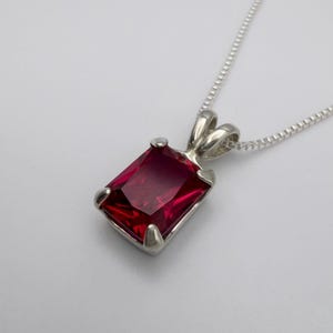 May include: A silver pendant necklace featuring a rectangular, deep red gemstone. The gemstone is set in a silver bezel with four prongs. The chain is a delicate silver box chain, creating a classic and elegant look.