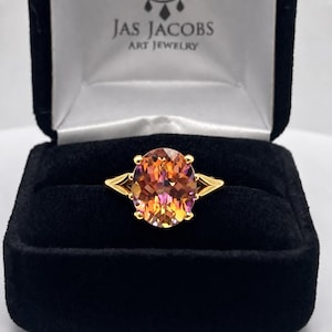 May include: A gold ring featuring a large, oval-cut, multi-colored gemstone with orange, pink, and yellow hues. The ring is displayed in a black velvet jewelry box with the text JAS JACOBS ART JEWELRY printed in white on the inner lid.