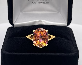 May include: A gold ring featuring a large, oval-cut, multi-colored gemstone with orange, pink, and yellow hues. The ring is displayed in a black velvet jewelry box with the text JAS JACOBS ART JEWELRY printed in white on the inner lid.
