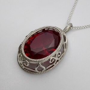 May include: Silver pendant necklace featuring an oval, faceted, deep red gemstone. The gemstone is set in a decorative, openwork silver frame with scroll designs. The necklace has a delicate silver chain.