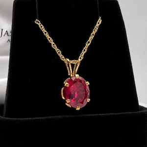 May include: A gold necklace featuring an oval-cut, deep red gemstone pendant held by a six-prong setting. The pendant is suspended from a delicate gold chain, showcasing a faceted surface that reflects light across the vibrant red stone.