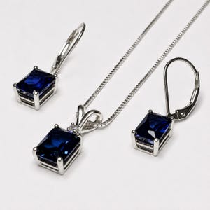 May include: A jewelry set consisting of a silver-toned necklace with a rectangular deep blue gemstone pendant and a pair of matching lever-back earrings, each featuring a single rectangular deep blue gemstone set in a silver-toned metal frame.