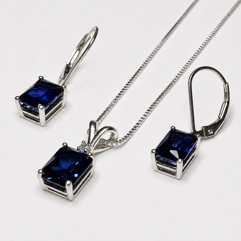 May include: A jewelry set consisting of a silver-toned necklace with a rectangular deep blue gemstone pendant and a pair of matching lever-back earrings, each featuring a single rectangular deep blue gemstone set in a silver-toned metal frame.
